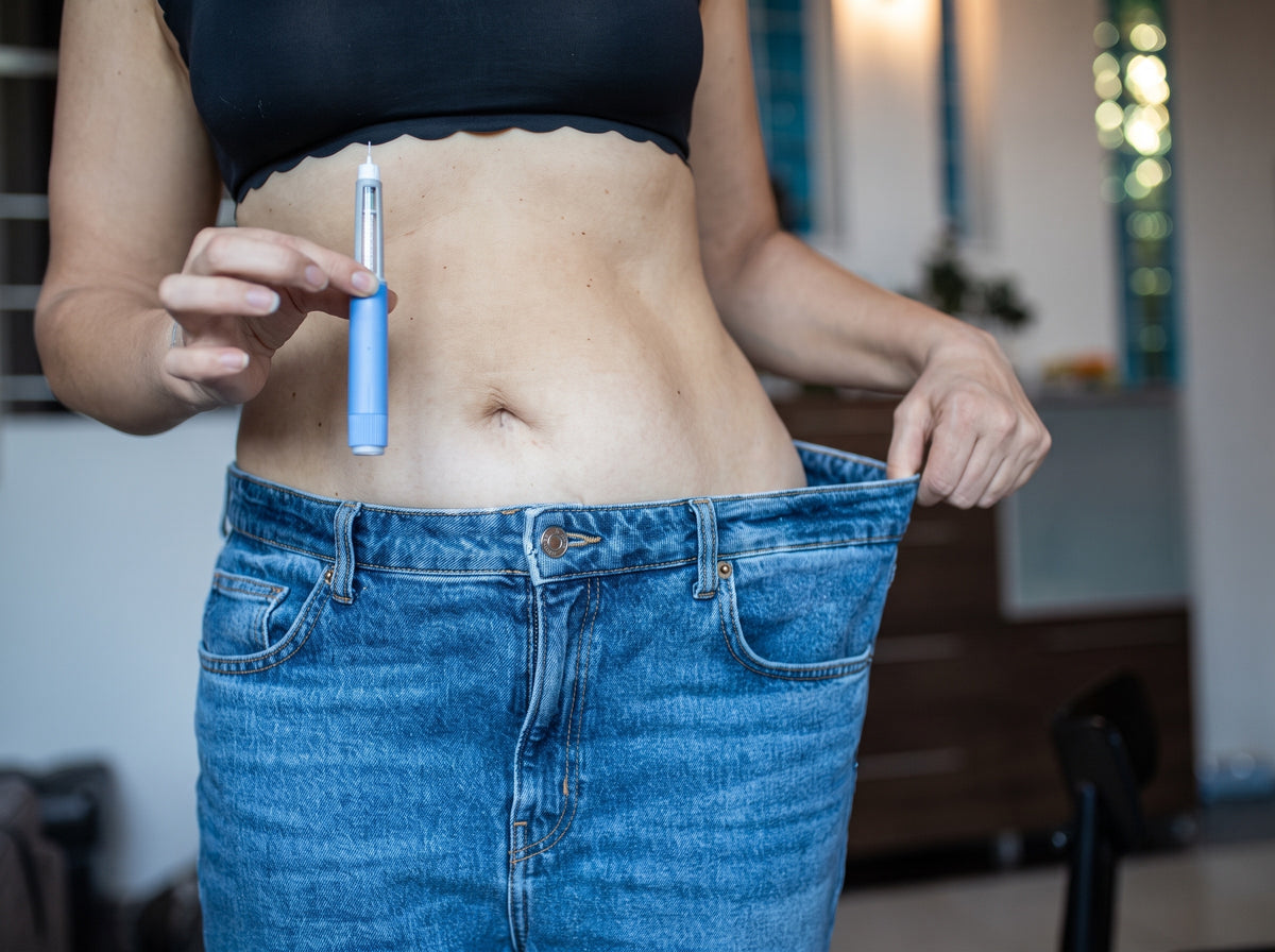 A woman holding the waistband of her old jeans out from her body with one hand, a GLP-1 pen in the other.