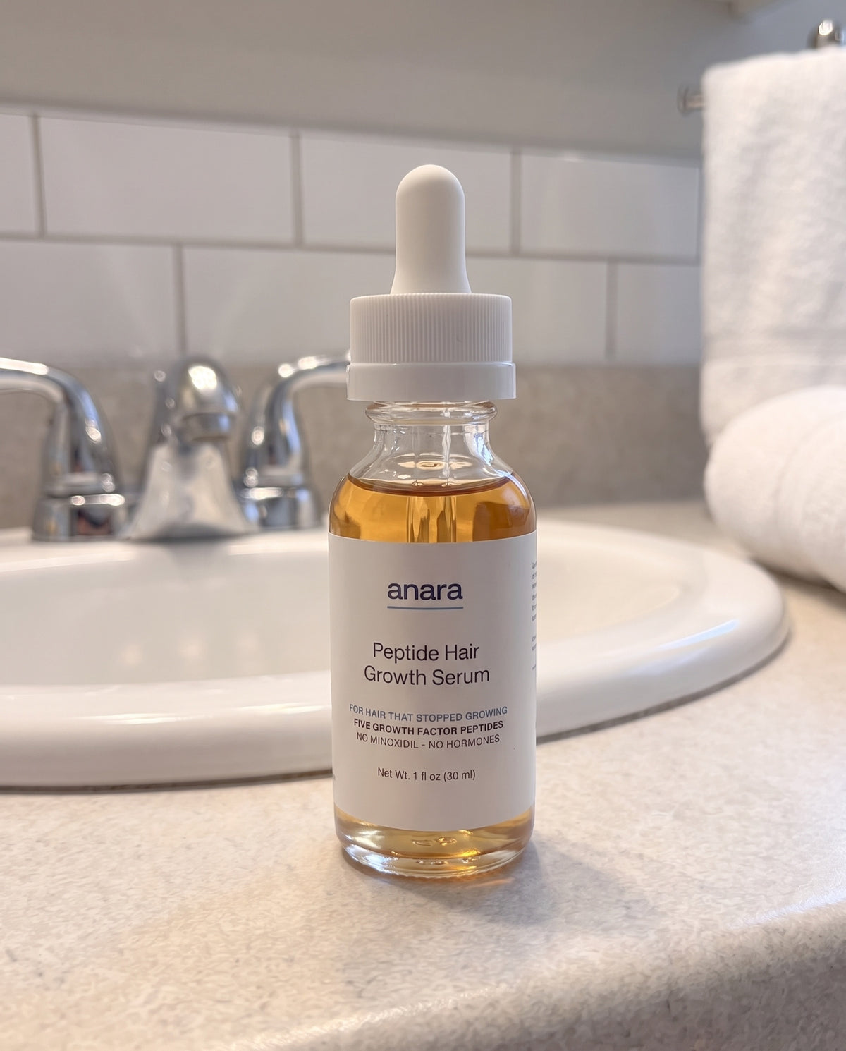 Anara Peptide Hair Growth Serum: a clear glass dropper bottle of amber serum with a white ribbed cap, standing on a bathroom sink counter in front of a chrome tap and a folded white towel.